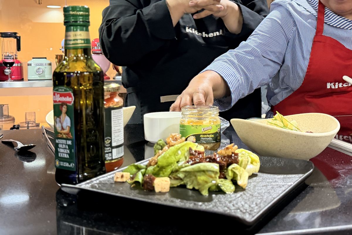 Mesa de trabajo con aceite de oliva La Española, un frasco de alcachofas Killio’s y un plato de ensalada.